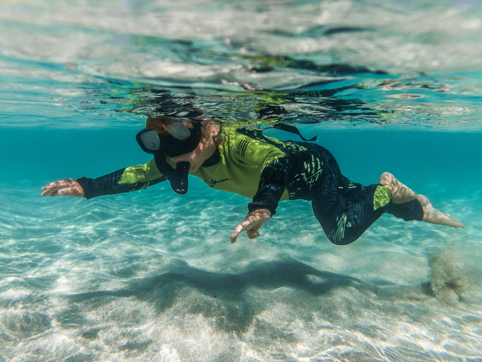 A child in a Waihana Lime Fishbone steamer wetsuit and black dive mask glides underwater through clear turquoise water above a sandy seafloor.