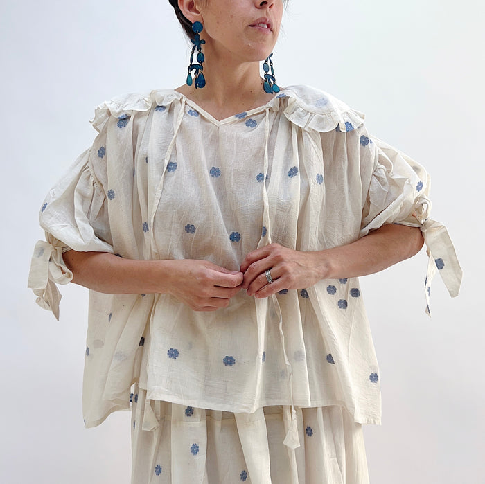 A woman wearing a white top with blue floral print with elbow length sleeves and a large collar with ruffle trim.
