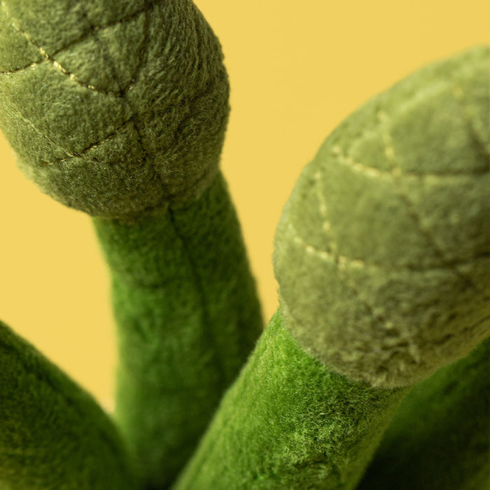 Extreme close-up of two Jellycat Amuseables Asparagus plush tips showing the dark green textured fabric and quilted cross-stitch detailing on the spear heads, against a yellow background