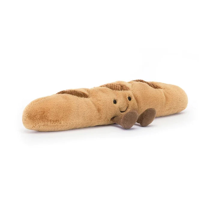 Jellycat Amuseables Baguette — a plush golden-brown French baguette character with black hard eyes, an embroidered smile, corduroy score marks on top, and small brown corduroy feet, lying on a white background