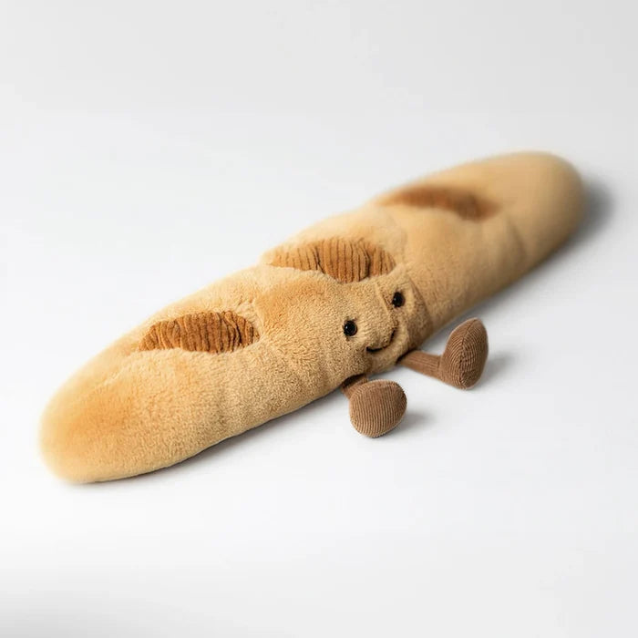 Jellycat Amuseables Baguette — three-quarter view of the plush baguette character showing its smiling face, corduroy score marks, and small brown feet, lying on a light grey background