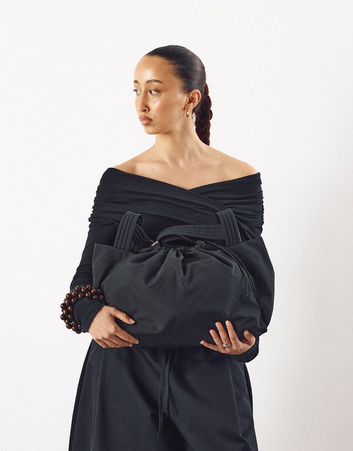 Woman wearing an off-shoulder black top holding a large black minimalist bag with gathered fabric and thick handles, showcasing understated elegance and modern design.