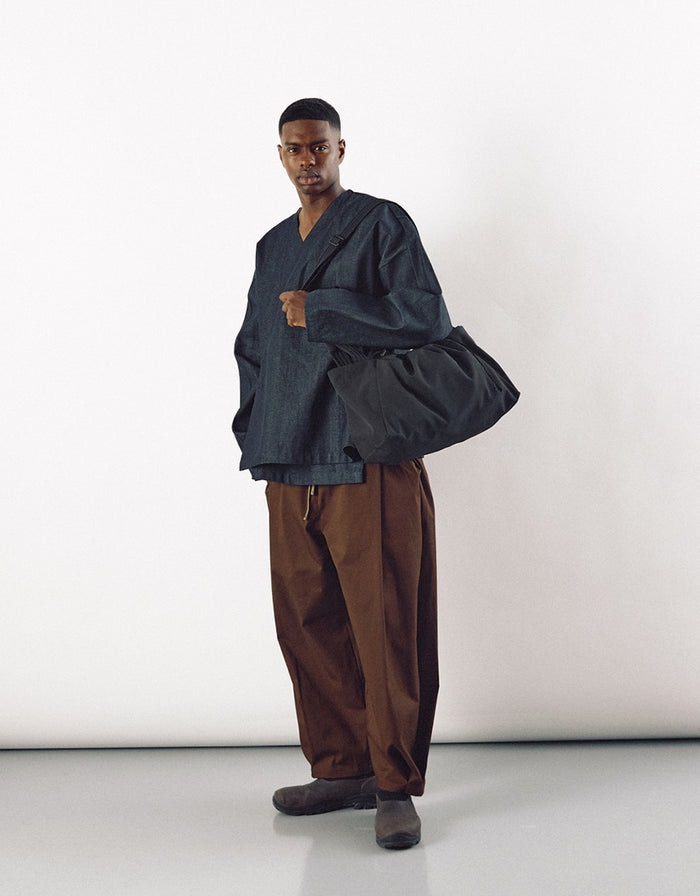 A man wearing a minimalist dark top and brown wide-leg pants, carrying a large black unisex bag that combines contemporary design with understated elegance.
