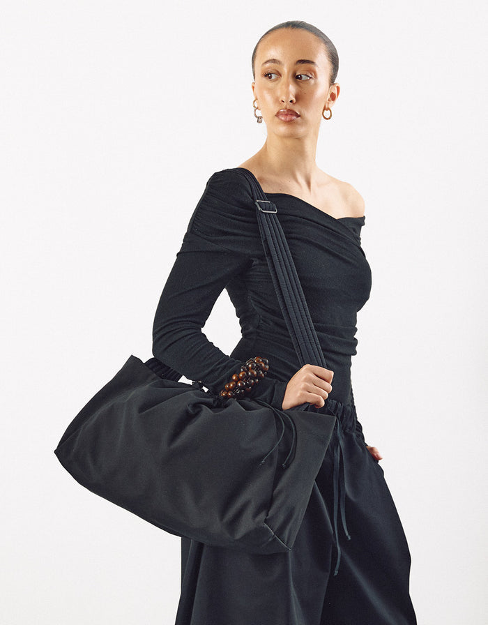 Woman wearing a minimalist black off-shoulder top and pants, carrying a large black maxi bag with a wide strap, highlighting understated elegance and volume.