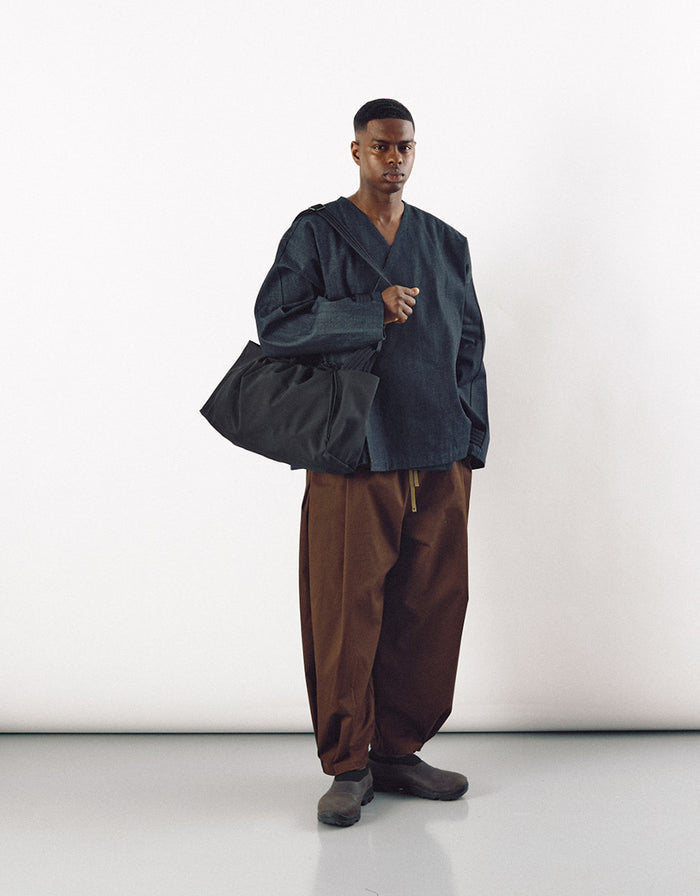 Male model wearing an oversized black top and brown wide-leg pants, carrying a large minimalist black bag designed for freedom of movement and understated elegance.