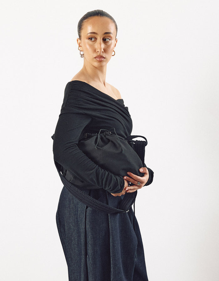 Woman wearing a minimalist off-shoulder black top holding a black compact bag with a drawstring closure, showcasing understated elegance and refined craftsmanship.