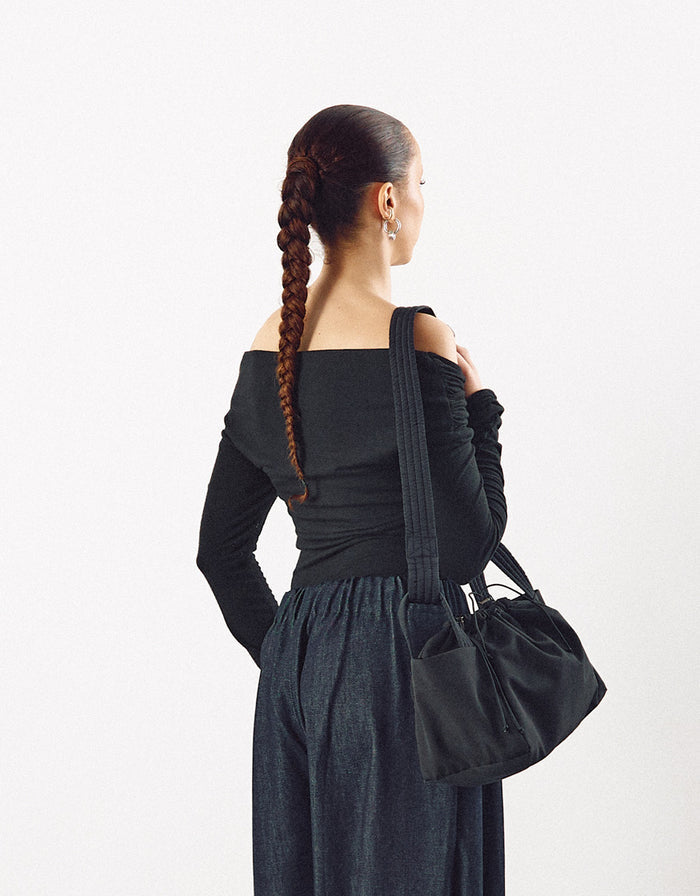 Woman seen from behind wearing a minimalist black off-shoulder top and loose dark pants, carrying a black structured shoulder bag with a modern, understated design.