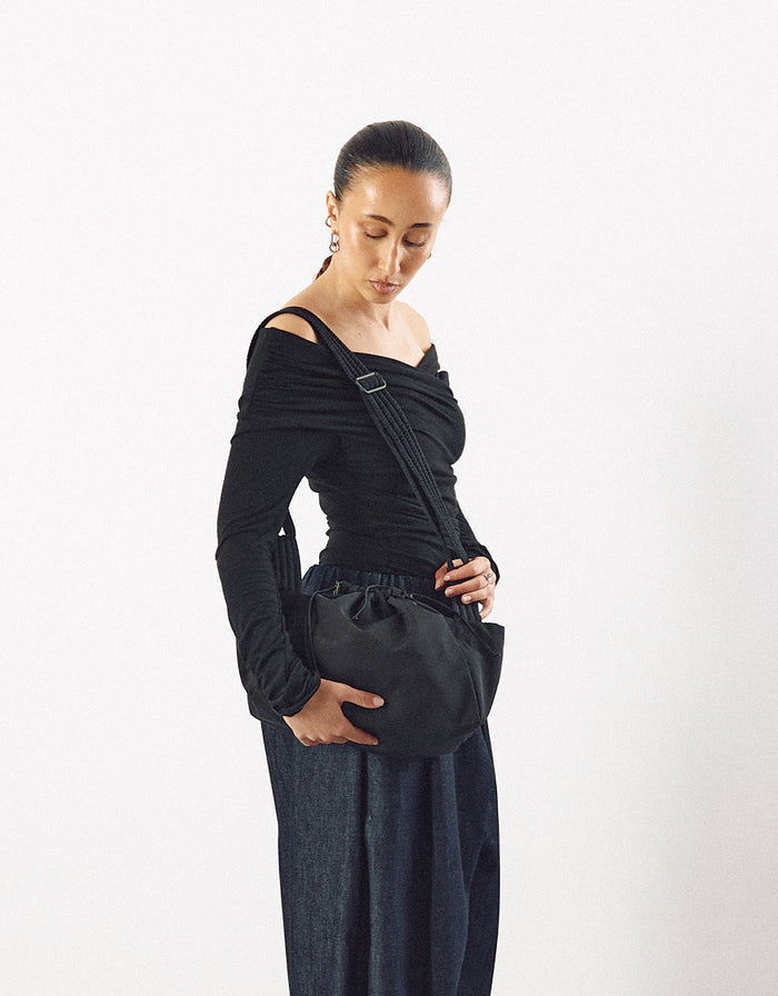 Woman wearing a black asymmetrical top and dark pants, carrying a minimalist black crossbody bag with a gathered detail and adjustable strap.