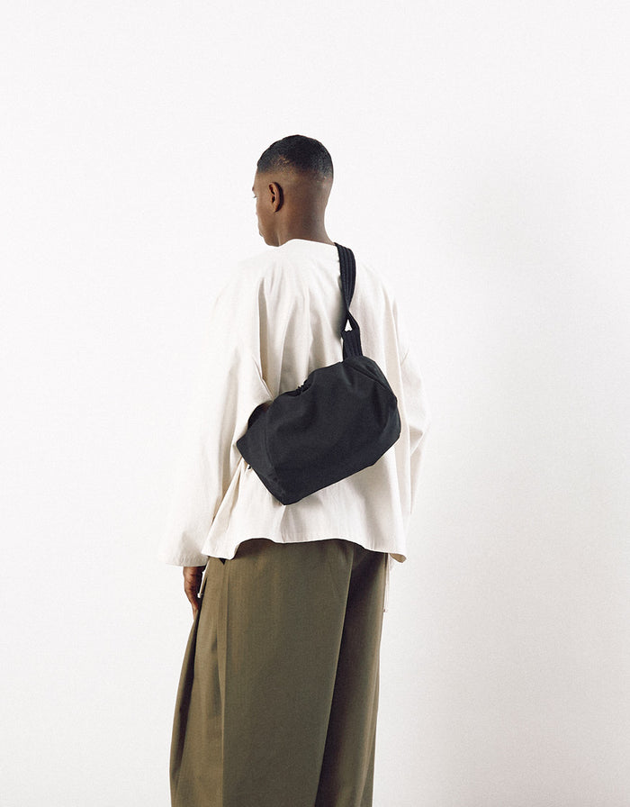 Man wearing a minimalist black bag with an adjustable strap across the back, paired with a loose white top and olive green wide-leg pants against a plain white background.