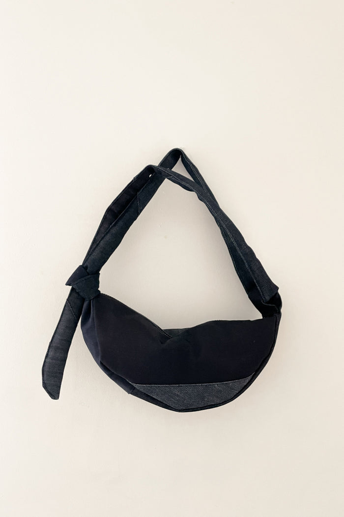 Minimalist unisex black denim shoulder bag with an asymmetrical design and adjustable knot detail, combining contemporary style with understated elegance.