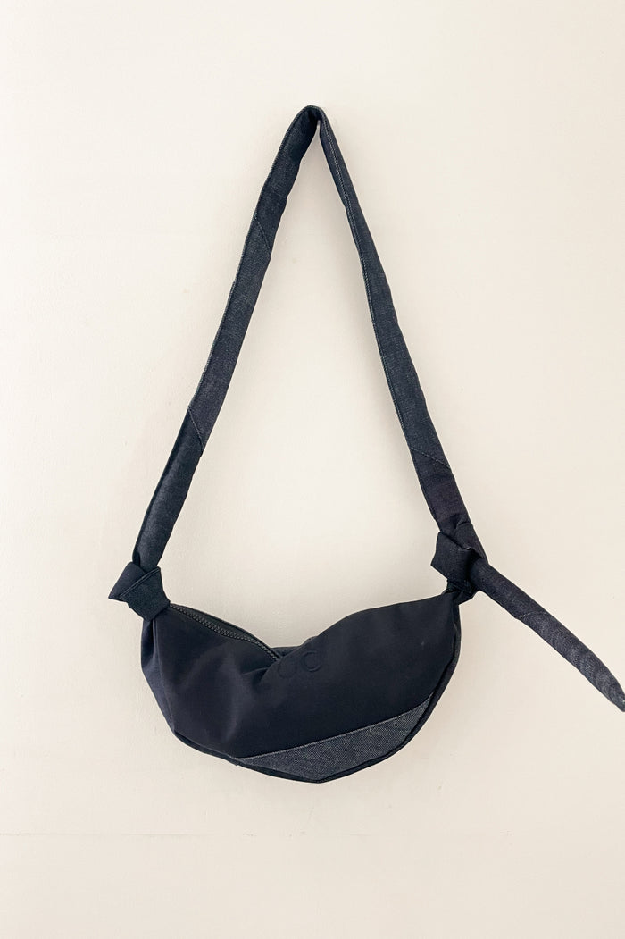 Minimalist unisex denim bag with an asymmetrical shape and long strap, showcasing refined craftsmanship and subtle design details in a deep blue tone.