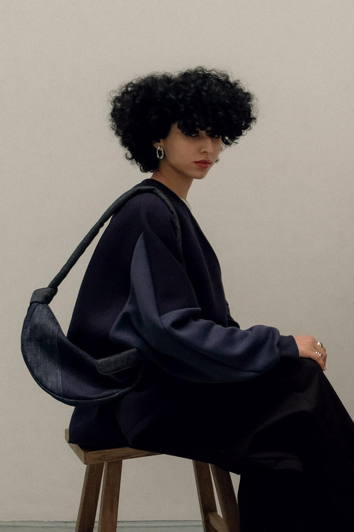 Woman with curly hair sitting on a wooden stool wearing a dark oversized top and carrying a minimalist denim bag with an asymmetrical design over her shoulder.
