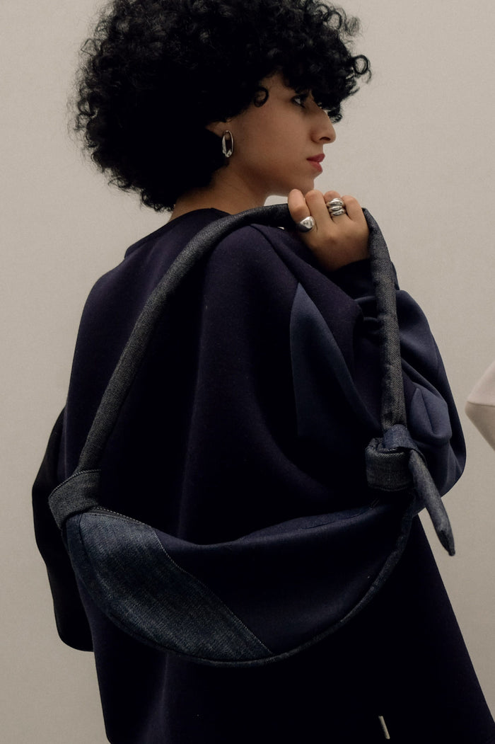 Woman with curly hair wearing silver earrings and rings, carrying a minimalist denim shoulder bag with an asymmetrical design over a dark, oversized garment.