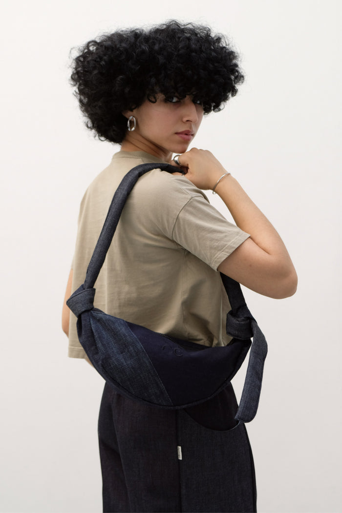 Woman wearing a casual beige t-shirt and dark denim pants, carrying a minimalist dark denim shoulder bag with knotted straps, showcasing understated elegance and contemporary design.