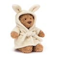 Jellycat Bartholomew Bear wearing a cream plush bathrobe with a bunny-eared hood pulled up, showing golden-brown curly fur face, black bead eyes, and brown nose, seated on a white background
