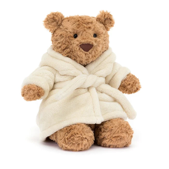 Jellycat Bartholomew Bear wearing a cream plush bathrobe with the hood down, showing golden-brown curly fur, round ears, black bead eyes, and brown nose, seated on a white background