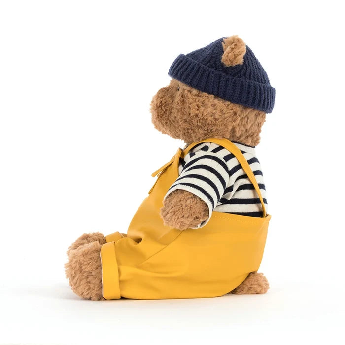 Side profile of Jellycat Bartholomew Bear in the seafarer outfit showing the navy beanie, striped top, mustard dungaree strap, and curly golden-brown fur, on a white background