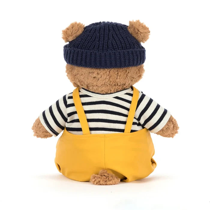 Rear view of Jellycat Bartholomew Bear in the seafarer outfit showing the navy beanie with two ear holes, striped top, mustard dungaree straps crossing the back, and a small curly tail, on a white background