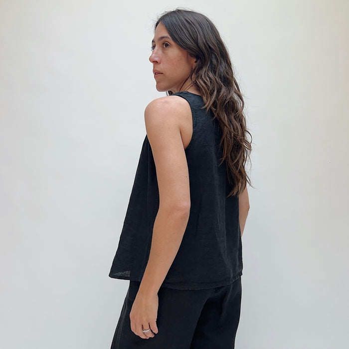 A person with long brown hair wearing a black linen tank top and matching black linen pants. She is turned away from the camera and looking over her shoulder.