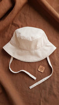 A flat lay photograph of a handmade baby bucket hat featuring fine beige and white vertical stripes. The hat includes matching fabric chin straps and is styled on a rich terracotta-colored linen backdrop next to a small square wooden compass charm.