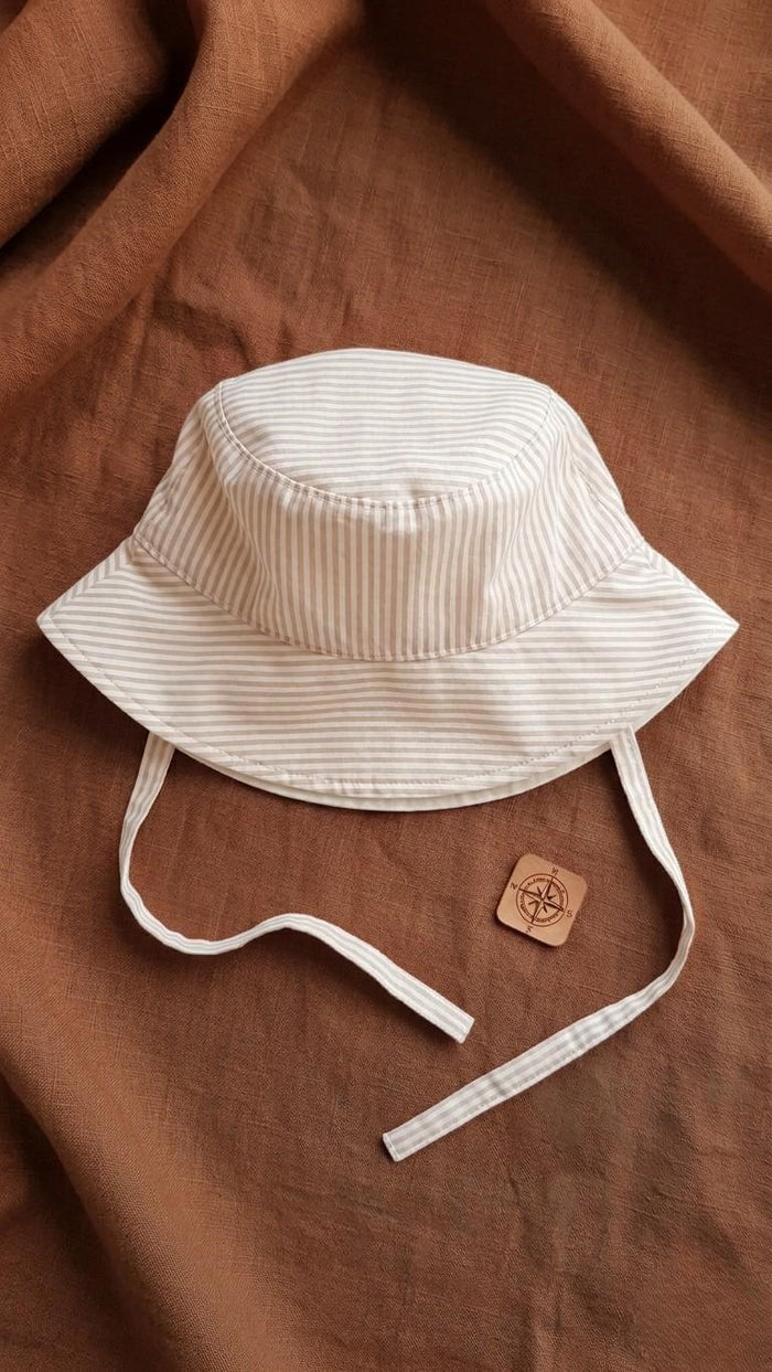 A flat lay photograph of a handmade baby bucket hat featuring fine beige and white vertical stripes. The hat includes matching fabric chin straps and is styled on a rich terracotta-colored linen backdrop next to a small square wooden compass charm.