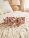 A soft pink muslin cotton baby headband featuring a dainty yellow, green, and purple floral pattern, finished with a matching fabric bow, displayed on a textured white cable-knit blanket.