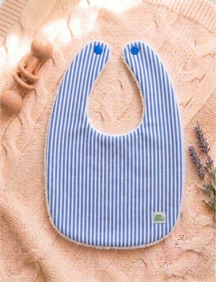 A flat lay photograph of a handmade baby bib featuring a blue and white vertical stripe pattern on the front and a white towelling texture on the back. The bib has two blue snap fasteners at the neck and a small green turtle brand label in the corner. It is styled on a soft, peach-colored cable knit blanket next to a wooden baby rattle and a sprig of dried lavender.