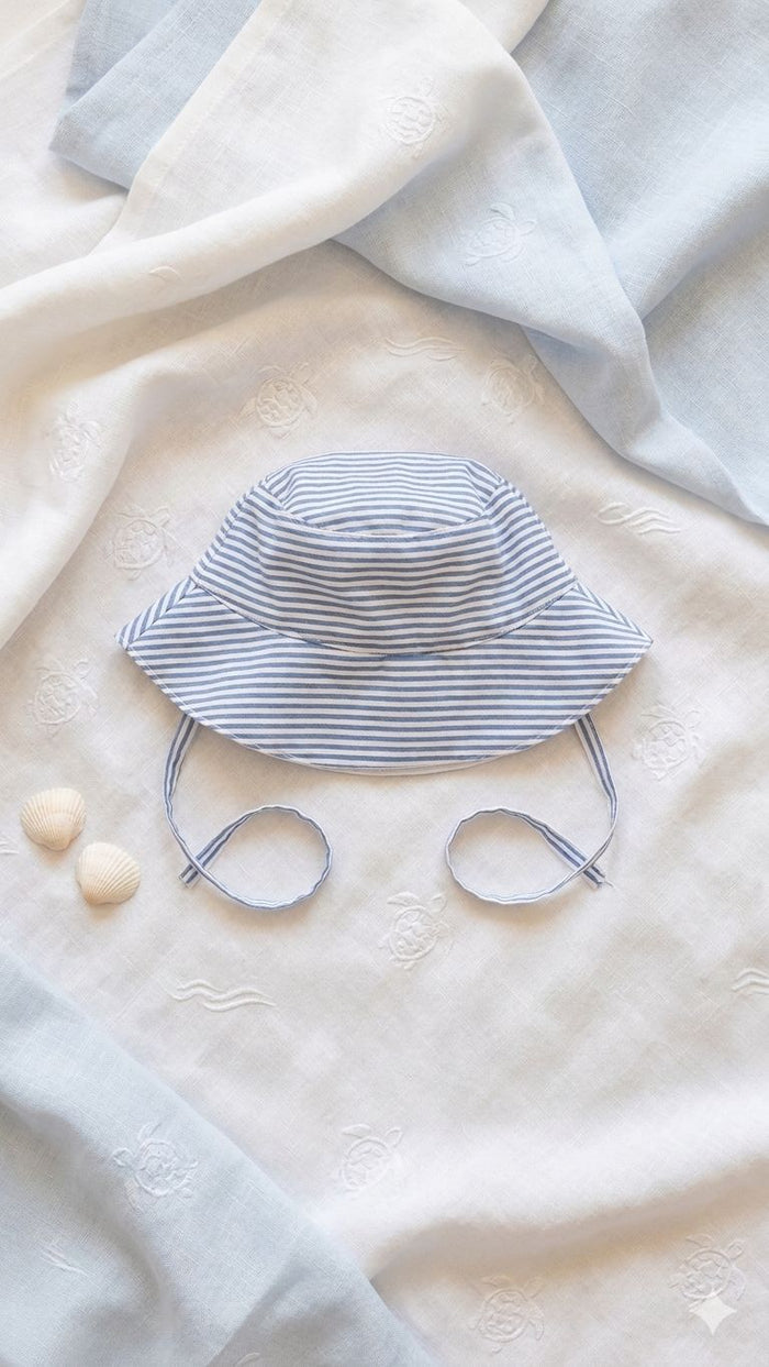 A flat lay photograph of a handmade baby bucket hat featuring blue and white horizontal stripes. The hat includes matching fabric chin straps and a small green turtle brand label on the brim. It is styled on a neutral linen backdrop alongside a sprig of dried lavender and a small smooth stone in soft, natural light.
