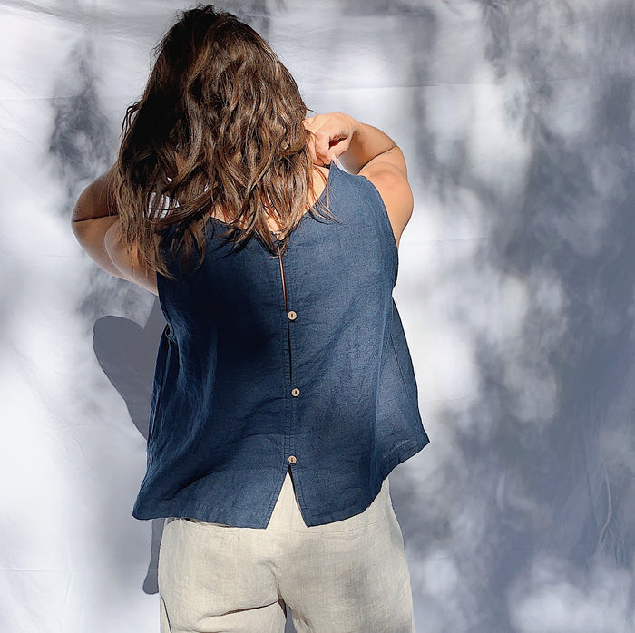 A person wearing a navy linen tank top with buttons down the back and side slits, as seen from behind with their hands raised to their shoulders. They have brown wavy hair.