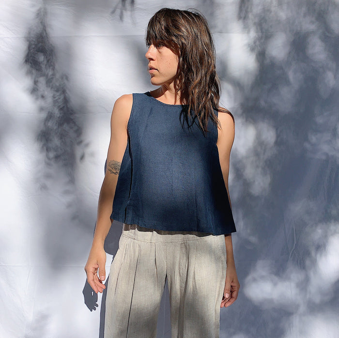 A person wearing a navy linen tank top with buttons down the back and side slits, looking off camera to the left, with their hands at their side. They have brown wavy hair.