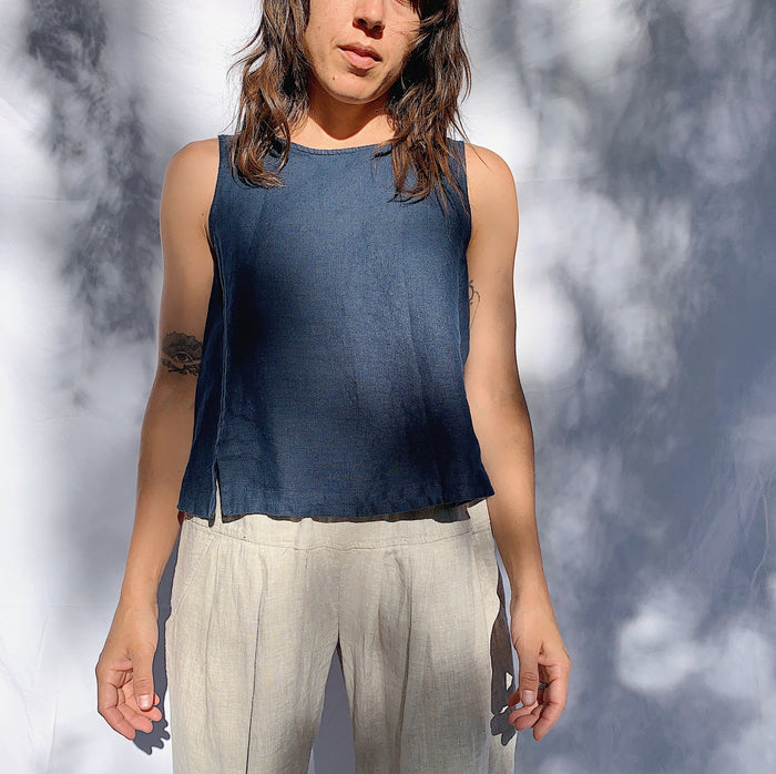 A person wearing a navy linen tank top with buttons down the back and side slits, looking off camera to the left, with their hands at their side. They have brown wavy hair.