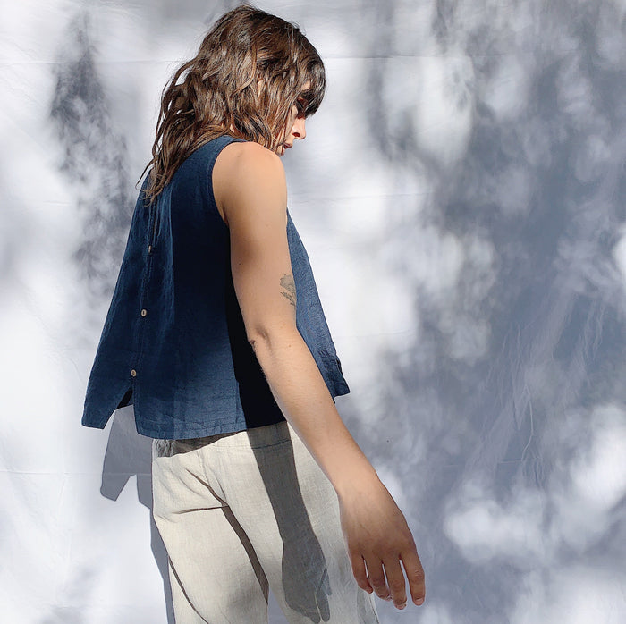 A person wearing a navy linen tank top with buttons down the back and side slits, turned half way around with their hands at their side. They have brown wavy hair.