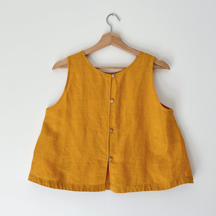 Mustard yellow sleeveless tank top on a wooden hanger against a white background as seen from the backside with buttons running down the tank top.