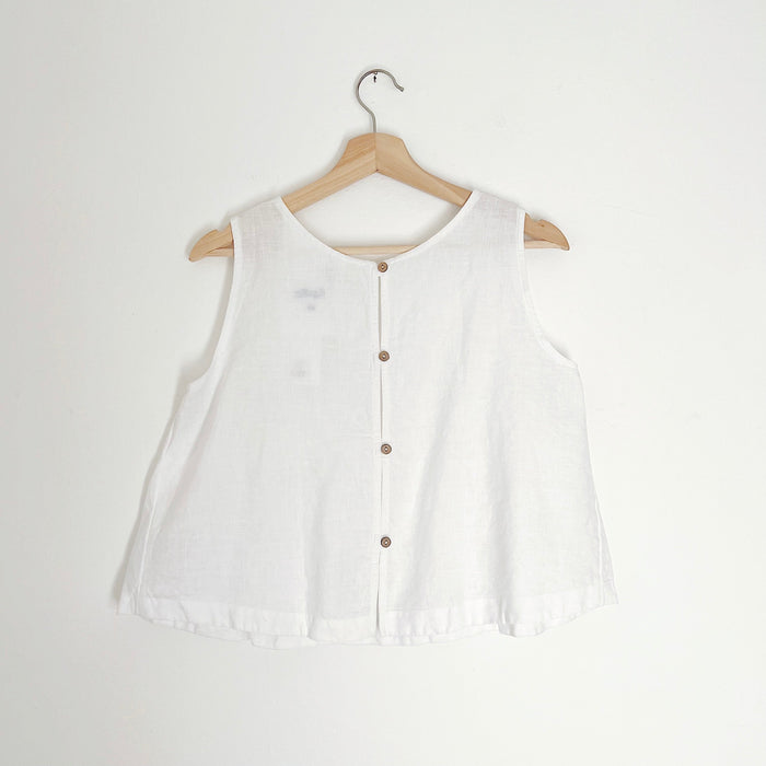 Cropped white linen tank top with buttons down the back and side slits, displayed on a wooden hanger against a white background.