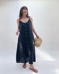 Woman with brown hair wearing a long black linen dress with buttons down the front, holding a tan straw hat on a white background.