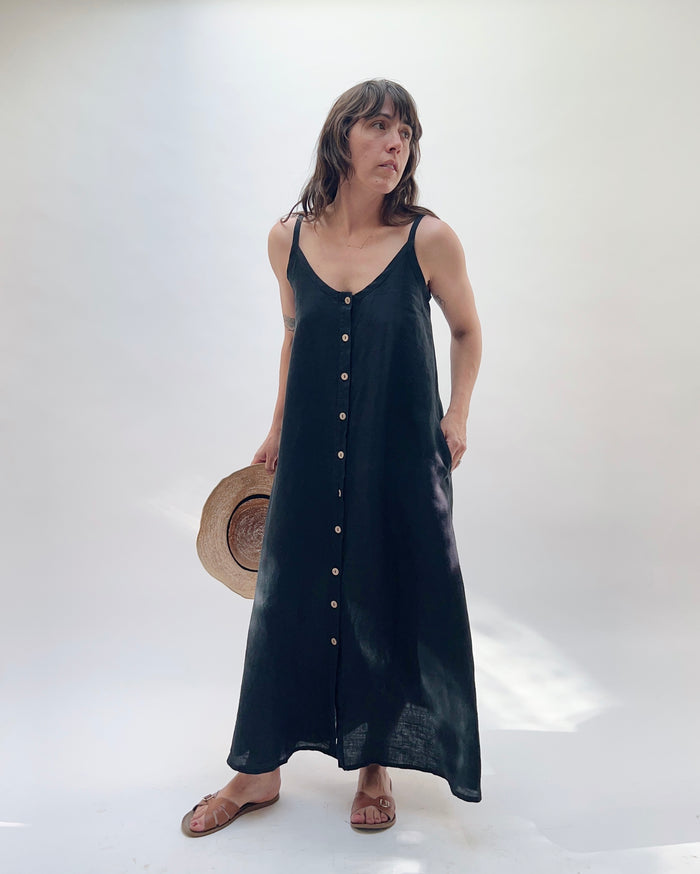 Woman with brown hair wearing a long black linen dress with buttons down the front, holding a tan straw hat on a white background.
