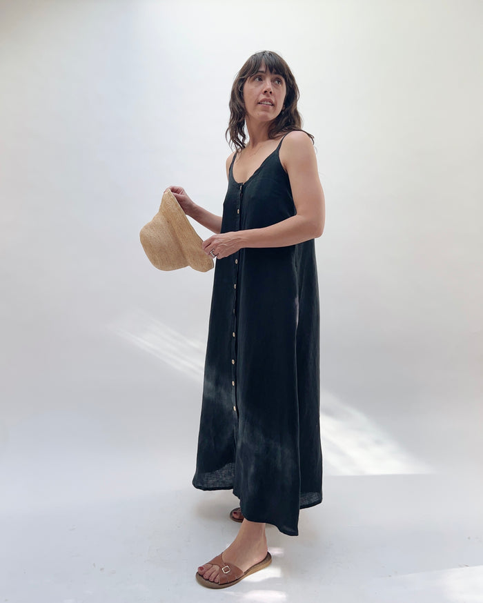 Woman with brown hair wearing a long black linen dress with buttons down the front, holding a tan straw hat on a white background turned to the side and looking out of frame.