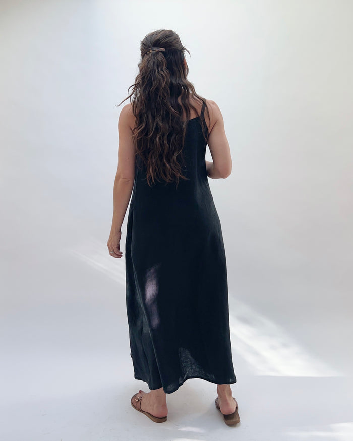 Woman with brown hair wearing a long black linen dress with buttons down the front on a white background as seen from behind.