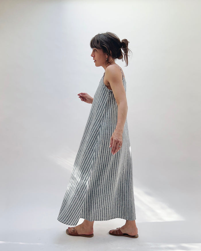 Woman wearing a long, striped linen dress in white and grey on a white background as seen from the side.