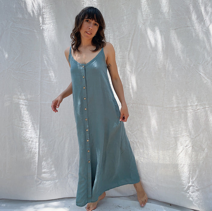 Woman with brown hair wearing a long sage green linen dress with buttons down the front on a canvas background.
