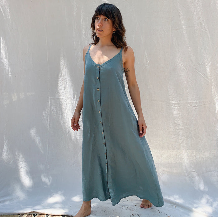 Woman with brown hair wearing a long sage green linen dress with buttons down the front on a canvas background.