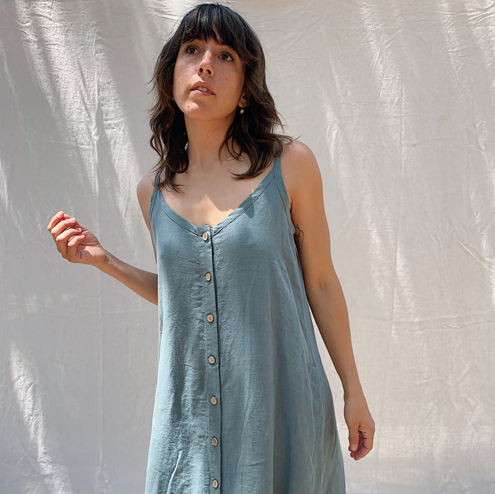 Close up shot of a woman with brown hair wearing a long sage green linen dress with buttons down the front on a canvas background.