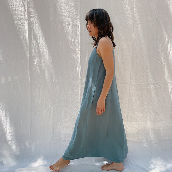 A woman seen from the side with brown hair wearing a long sage green linen dress with buttons down the front on a canvas background.