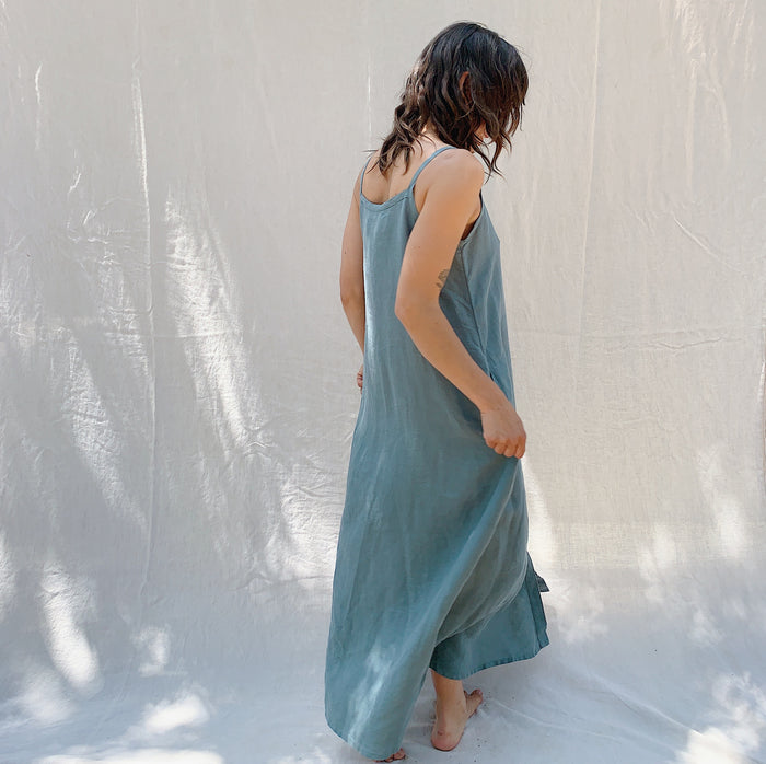 A woman seen from behind with brown hair wearing a long sage green linen dress with buttons down the front on a canvas background.