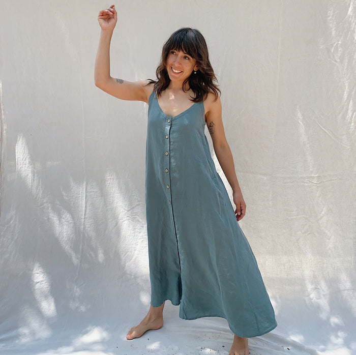 A woman holding up one arm in the air with brown hair wearing a long sage green linen dress with buttons down the front on a canvas background.