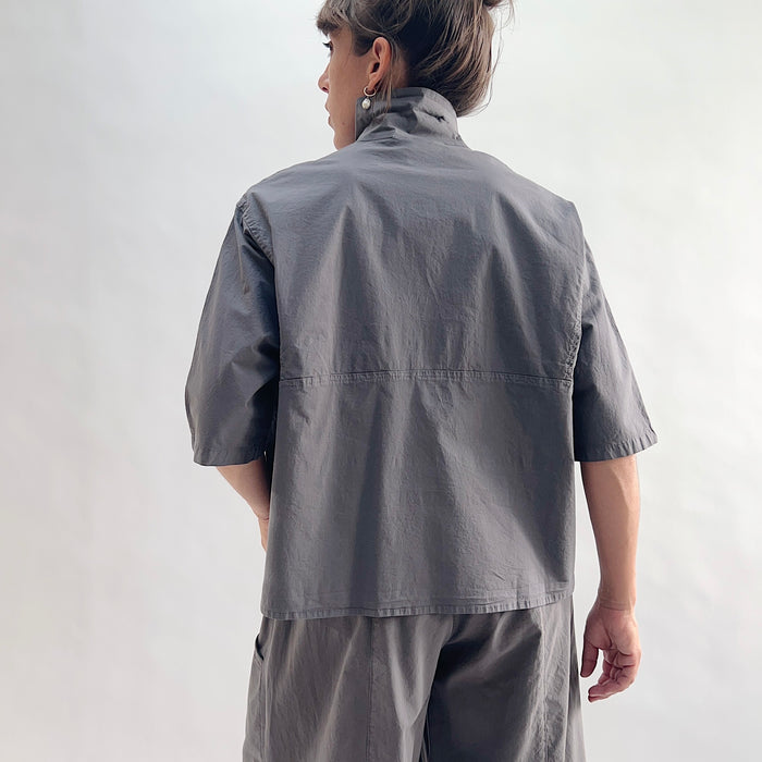 Back view of model wearing Eleven Stitch eiffel tower camp shirt, showing point collar, back yoke seam, short sleeves with rolled cuffs, slightly cropped boxy silhouette, and deep slate grey color, styled with matching eiffel tower one pocket pant