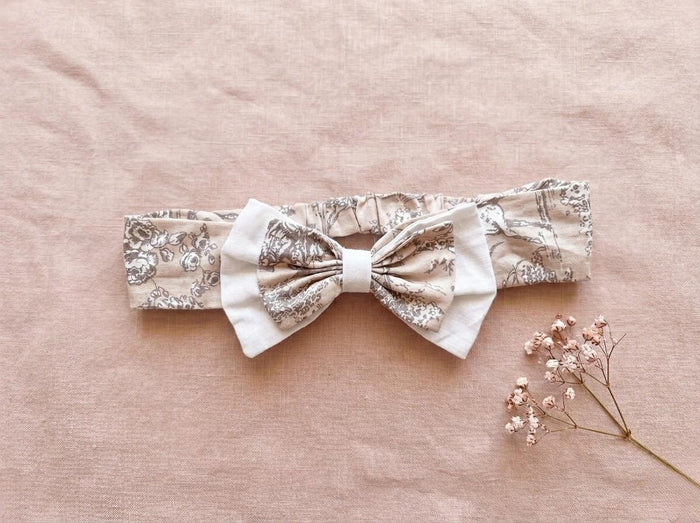 A flat lay photograph of the Carmen baby headband, featuring a beige fabric with a delicate brown vintage floral or toile-style pattern. The headband includes an elasticated back and is adorned with a layered bow—a smaller floral bow set atop a larger white fabric bow. It is styled against a soft, dusty pink linen backdrop next to a sprig of dried pink baby's breath flowers.