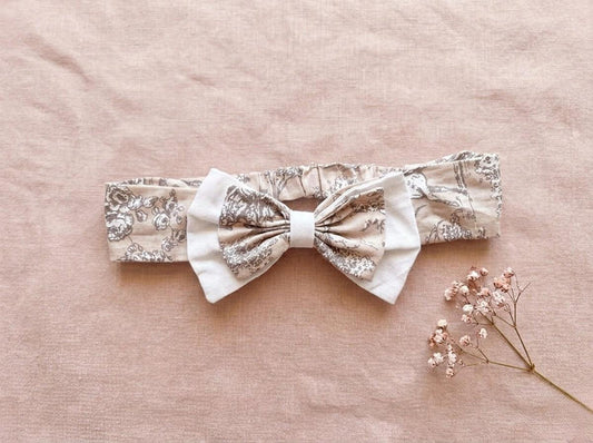 A flat lay photograph of the Carmen baby headband, featuring a beige fabric with a delicate brown vintage floral or toile-style pattern. The headband includes an elasticated back and is adorned with a layered bow—a smaller floral bow set atop a larger white fabric bow. It is styled against a soft, dusty pink linen backdrop next to a sprig of dried pink baby's breath flowers.