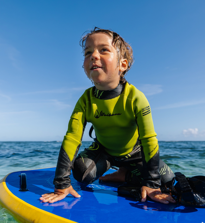 A happy young child in a Waihana lime green and black Fishbone Steamer full wetsuit sits on a blue surfboard on calm ocean water under a bright blue sky.
