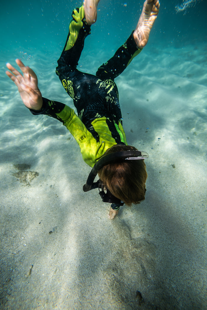 A child in a Waihana Lime Fishbone Steamer wetsuit dives toward a sandy ocean floor in clear turquoise water, wearing a black mask and snorkel.
